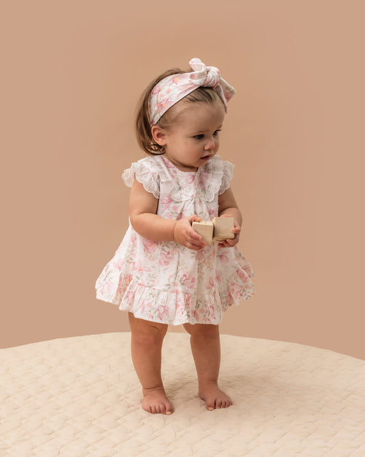 This gorgeous floral print over romper dress features shoulder frills with broderie lace frill on yoke seam, hem frill, back neck button opening, and a press stud crotch opening for ease of dressing.